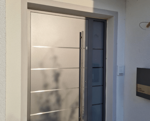 Helle Aluminium-Haustür mit Edelstahl-Zierleisten und Stoßgriff neben anthrazitfarbenem Seitenteil